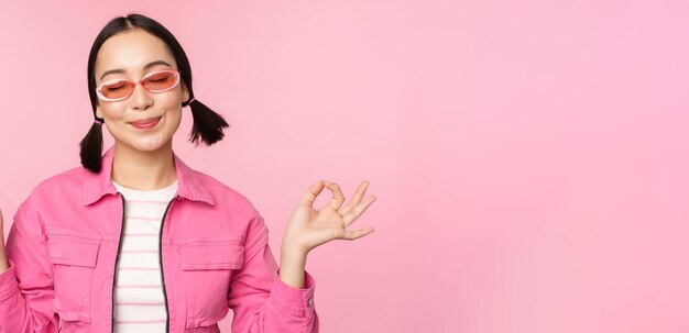Achtsamkeits- und Wellness-Konzept Lächelndes koreanisches Mädchen in stilvollem Outfit, das das Hörmantra meditiert, hält die Hände in Zen-Peace-Pose und praktiziert Yoga, das über rosafarbenem Hintergrund steht