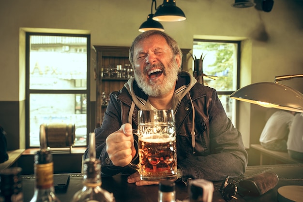 Kostenloses Foto Älterer bärtiger mann, der alkohol in der kneipe trinkt und ein sportprogramm im fernsehen sieht. ich genieße meinen lieblingsbrot und mein lieblingsbier. mann mit bierkrug, der am tisch sitzt. fußball- oder sportfan. konzept der menschlichen emotionen