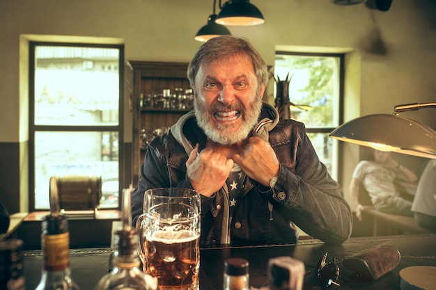 Kostenloses Foto Älterer bärtiger mann, der alkohol in der kneipe trinkt und ein sportprogramm im fernsehen sieht. ich genieße meinen lieblingsbrot und mein lieblingsbier. mann mit bierkrug, der am tisch sitzt. fußball- oder sportfan. konzept der menschlichen emotionen