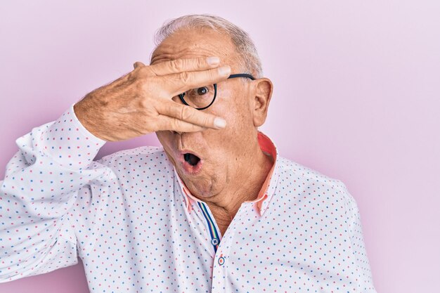 Älterer kaukasischer mann, der lässige kleidung und brille trägt und schockiert späht, gesicht und augen mit der hand bedeckt und ängstlich durch die finger schaut