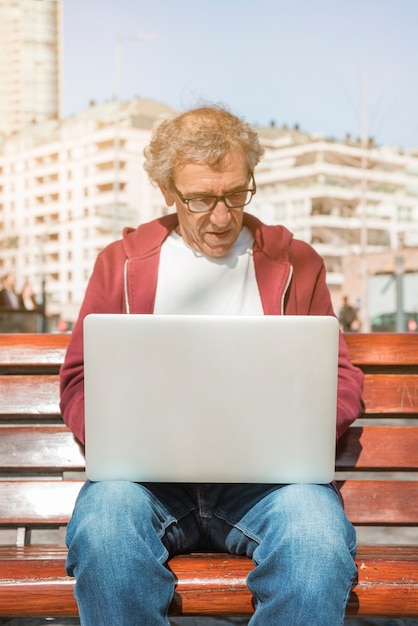 Kostenloses Foto Älterer mann, der auf bank unter verwendung des laptops an draußen sitzt