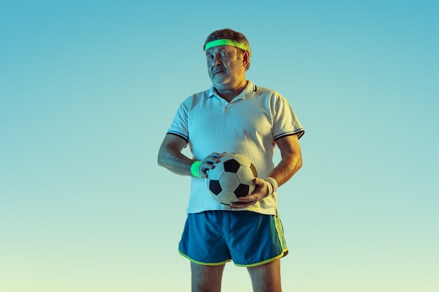 Älterer mann, der fußball in sportbekleidung auf gradientenhintergrund und neonlicht spielt