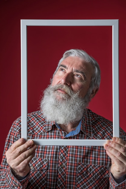 Kostenloses Foto Älterer mann, der oben schaut, weißen grenzrahmen vor seinem gesicht gegen roten hintergrund halten