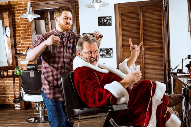 älterer Mann im Weihnachtsmannkostüm, das seinen persönlichen Meister im Friseurladen vor Weihnachten rasiert