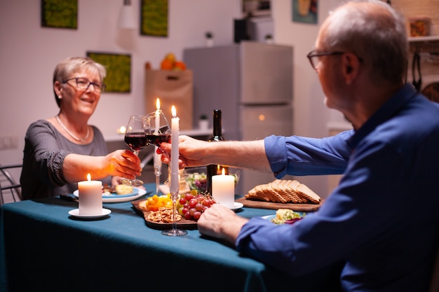 Älteres paar, das weingläser während der beziehungsfeier in der küche am abend hält. älteres ehepaar sitzt am tisch im esszimmer, redet, genießt das essen,