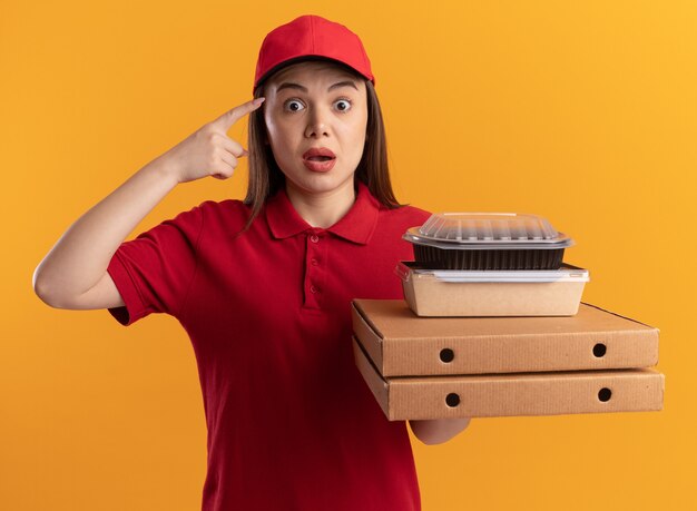 Ängstliche hübsche lieferfrau in einheitlichen punkten am kopf und hält papiernahrungsmittelpakete auf pizzaschachteln auf orange