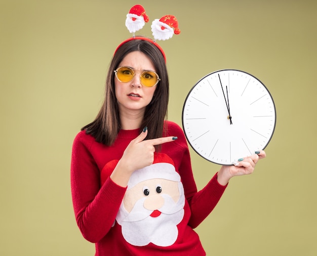 Kostenloses Foto Ängstliche junge hübsche kaukasische mädchen mit weihnachtsmann-pullover und stirnband mit brille, die die uhr hält und auf die kamera zeigt, die auf olivgrünem hintergrund isoliert ist