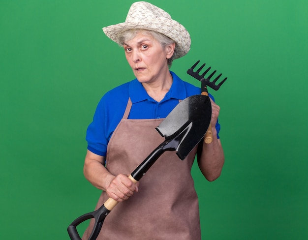 Kostenloses Foto Ängstlicher älterer weiblicher gärtner, der gartenhut hält, der spaten und rechen auf grün hält