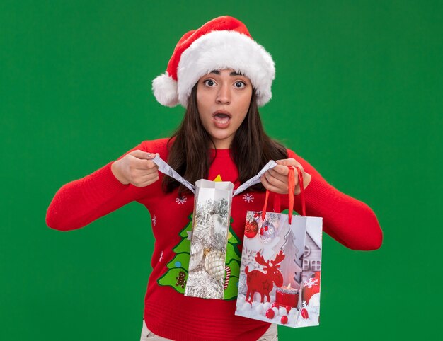 ängstliches junges kaukasisches Mädchen mit Weihnachtsmütze hält Papiergeschenktüten lokalisiert auf grünem Hintergrund mit Kopienraum