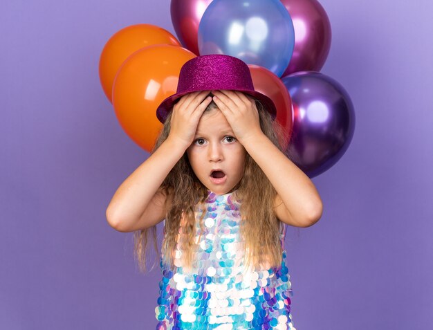 Ängstliches kleines blondes mädchen mit violettem partyhut, das die hände auf die stirn legt und mit heliumballons isoliert auf lila wand mit kopierraum steht