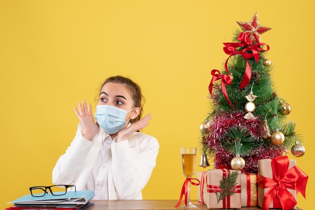 Ärztin der vorderansicht, die in der schutzmaske sitzt, erschrocken auf gelbem hintergrund mit weihnachtsbaum und geschenkboxen