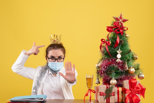 Ärztin der vorderansicht, die in der sterilen maske auf gelbem hintergrund mit weihnachtsbaum und geschenkboxen sitzt