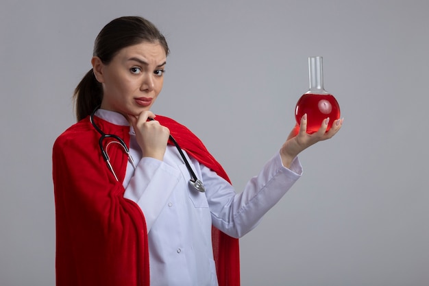 Ärztin mit stethoskop in weißer medizinischer uniform und rotem superheldenumhang, der kolben mit roter flüssigkeit hält, die vorne mit nachdenklichem ausdruck betrachtet, der über weiße wand steht