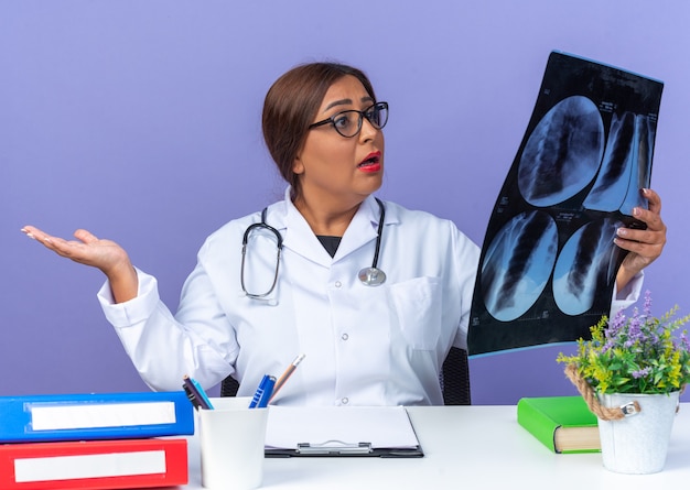 Ärztin mittleren alters im weißen kittel mit stethoskop mit brille, die eine röntgenaufnahme hält, die mit ausgestrecktem arm am tisch auf blauem hintergrund verwechselt wird