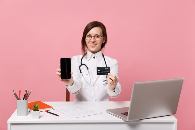 Ärztin sitzt am schreibtisch am computer mit medizinischem dokument halten handy im krankenhaus isoliert auf pastellrosa hintergrund. frau im medizinischen kittelbrillenstethoskop. konzept der gesundheitsmedizin.