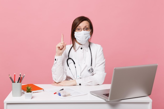 Ärztin sitzt an der schreibtischarbeit am computer mit medizinischem dokument in gesichtsmaske im krankenhaus einzeln auf pastellrosa wandhintergrund. frau im medizinischen kittelbrillenstethoskop gesundheitswesen medizinkonzept