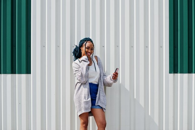 Afrikanerin mit Dreads-Haaren in Jeans-Shorts posiert mit Handy in der Hand vor weißer Stahlwand