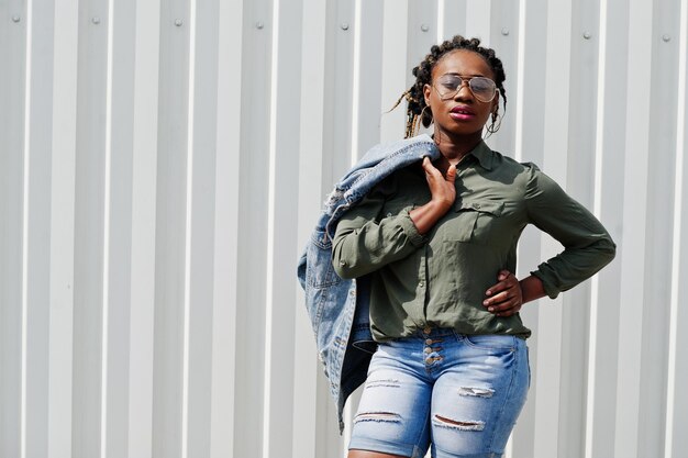 Afrikanerin mit Dreads-Haaren in Jeansjacke und Shorts trägt eine Brille, die vor einer weißen Stahlwand steht