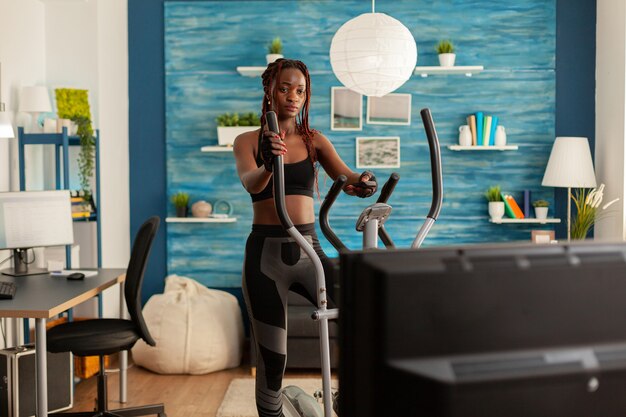 Afrikanische fitte starke Frau, die Cardio-Übungen auf einer Ellipsenmaschine macht, im Wohnzimmer zu Hause auf den Fernseher schaut und Anweisungen mit Fernbedienung sieht. Trainieren in Sportkleidung.