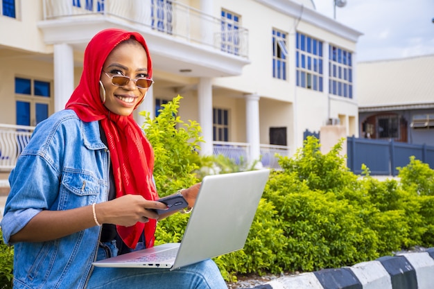 Afrikanische Frau, die draußen mit ihrem Laptop und ihrem Telefon sitzt und eine Online-Zahlung tätigt