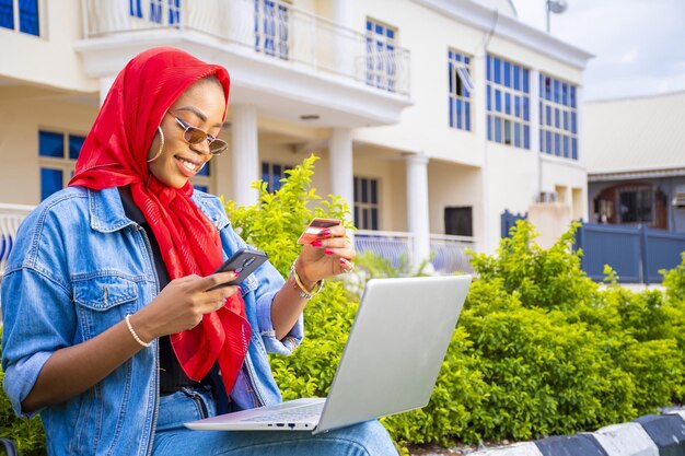 Afrikanische Frau, die draußen mit ihrem Laptop und ihrem Telefon sitzt und eine Online-Zahlung tätigt