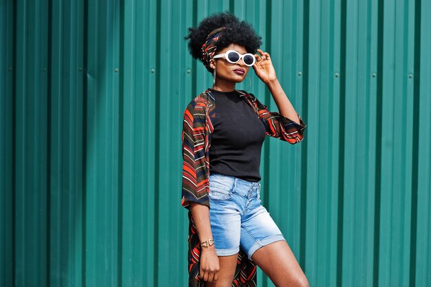 Afrikanische Frau mit Afro-Haaren in Jeans-Shorts und weißer Sonnenbrille posierte vor grüner Stahlwand