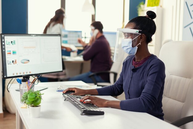 Afrikanische Frau tippt am Computer am Arbeitsplatz mit Gesichtsmaske als Sicherheitsvorkehrung gegen Covid19