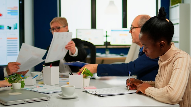 Afrikanische Geschäftsfrau, die Dokumente liest, unterschreibt, während Geschäftspartner Papierkram teilen, die am Konferenztisch im Broadroom sitzen. Vorstandssitzung mit Aktionären im Start-up-Büro