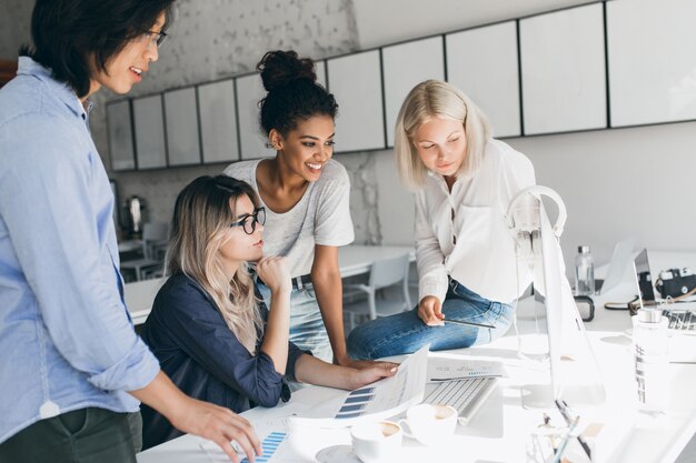 Afrikanische und europäische Mädchen, die Computerbildschirm betrachten, während blonde Dame Dokumente liest. Innenporträt des asiatischen Studenten in den Gläsern, die mit Freundinnen in der Einheit sprechen.