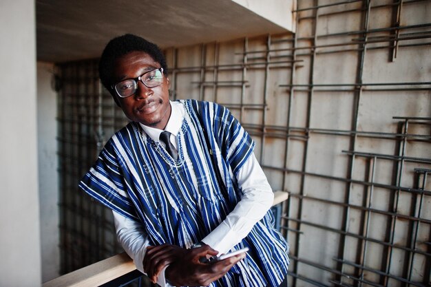 Afrikanischer Geschäftsmann in traditioneller Kleidung und Brille mit Handy zur Hand posiert in einem modernen Gebäude im Innenbereich