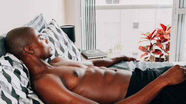 Afrikanischer junger Mann des hemdlosen Sitzes, der auf Bett sich entspannt