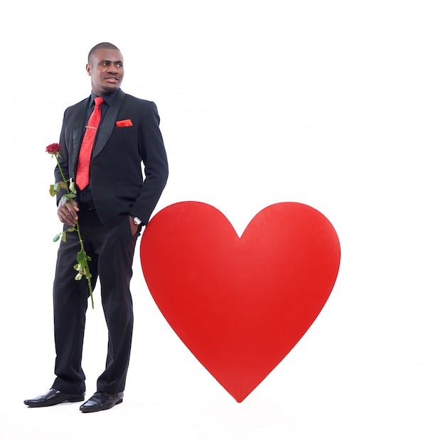 afrikanischer Mann, der vom großen verzierten roten Herzen lehnt und rote Rose hält