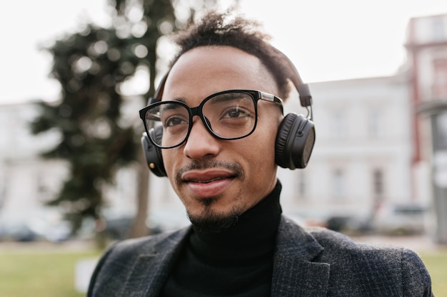 Afrikanischer mann in den gläsern, die auf stadt mit sanftem lächeln aufwerfen. wunderschöner brünetter schwarzer, der im freien in großen kopfhörern chillt.