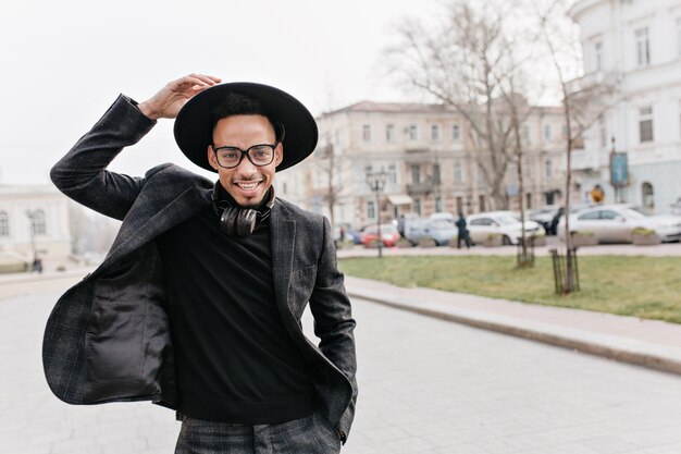 Afrikanischer Mann mit dem selbstbewussten Lächeln, das auf Unschärfestadt aufwirft. Foto im Freien des positiven großen Mulattenkerls, der auf der Straße lächelt.