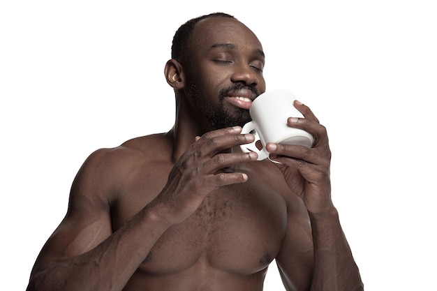Afrikanischer Mann mit weißer Tasse Tee oder Kaffee, lokalisiert auf weißem Studiohintergrund. Schließen Sie herauf Porträt im Minimalismusstil eines jungen nackten glücklichen Afro-Mannes