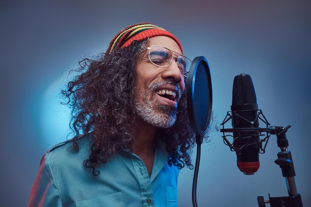 Afrikanischer Rastafari-Sänger, der ein blaues Hemd und eine Mütze trägt und im Aufnahmestudio emotional ein Lied schreibt. Isoliert auf blauem Hintergrund.