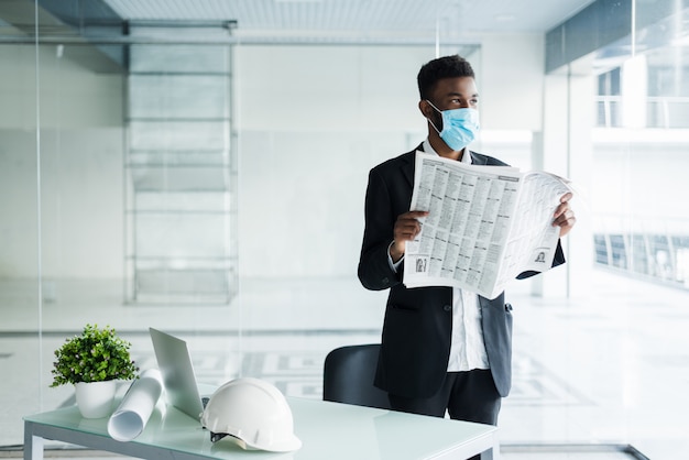 Afrikanischer schöner Geschäftsmann in der medizinischen Maske, die die Zeitung am Bürogebäude liest