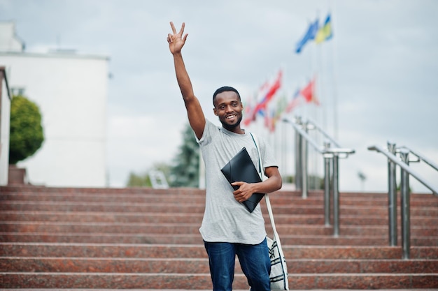 Afrikanischer Student männlich posiert mit Rucksack und Schulsachen auf dem Hof der Universität gegen Flaggen verschiedener Länder Zeigen Sie zwei Finger