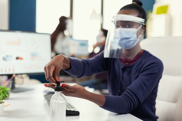 Afrikanischer Unternehmer, der am Arbeitsplatz Händedesinfektionsmittel mit Gesichtsmaske verwendet. Geschäftsfrau am neuen normalen Arbeitsplatz desinfiziert, während Kollegen im Hintergrund arbeiten.