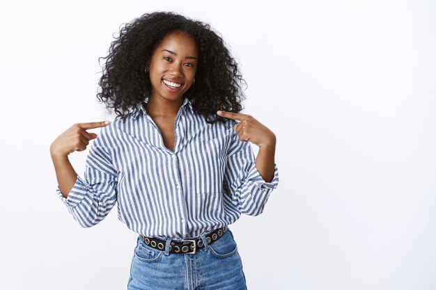 Afrikanisches Mädchen, das vorschlägt, die eigenen Fähigkeiten zu fördern, indem es sich selbst stolz lächelt, weiße Zähne, freundlich aussehender, geneigter Kopf, der selbstbewusst und ehrgeizig steht, Studiowand