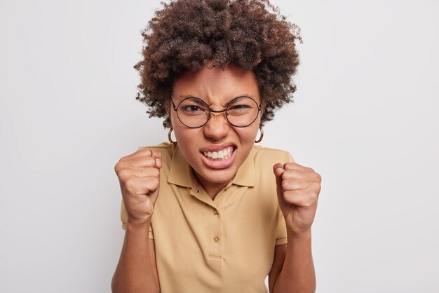 Afro-Amerikanerin beißt Zähne und Fäuste zusammen und ist wütend, weil sie von etwas gereizt ist, das störende Geräusche nicht ertragen kann, trägt ein lässiges T-Shirt mit Brille, das über einer grauen Wand isoliert ist