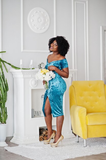 Afro-amerikanische Frauen in türkisfarbenem Kleid mit glänzenden Glitzerpailletten, goldenen Higheels, die mit Blumen vor einem weißen Raum mit gelbem Stuhl und Kamin stehen