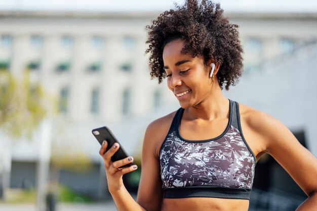 Afro sportliche Frau, die ihr Handy benutzt und sich nach dem Training im Freien entspannt