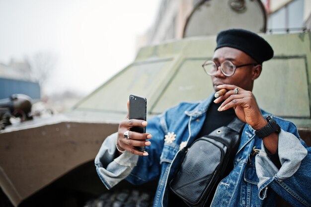 Afroamerikaner in Jeansjacke Baskenmütze und Brille raucht Zigarre und posiert gegen btr-Militärpanzer, der Selfie am Telefon macht