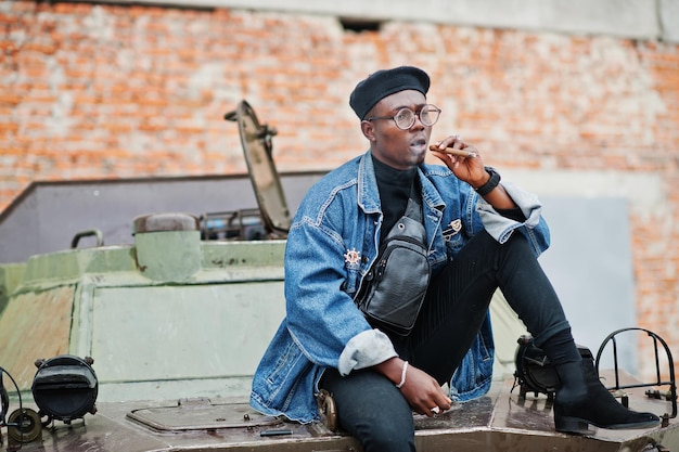 Kostenloses Foto afroamerikaner in jeansjacke baskenmütze und brille raucht zigarre und posiert gegen btr-militärpanzer