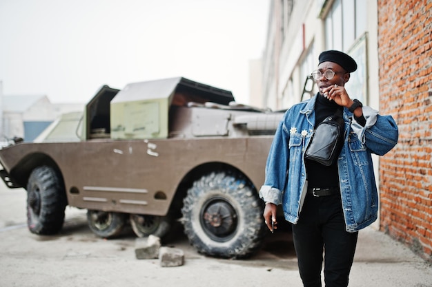 Afroamerikaner in Jeansjacke Baskenmütze und Brille raucht Zigarre und posiert gegen btr-Militärpanzer