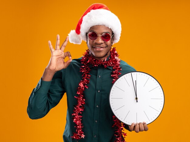 Afroamerikaner-Mann im Weihnachtsmannhut mit Girlande, die Sonnenbrille hält, die Uhr schaut, die Kamera lächelnd zeigt, ok Zeichen zeigend über orange Hintergrund