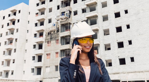 Afroamerikanerdame im Schutzhelm sprechend auf Smartphone nahe im Bau errichten