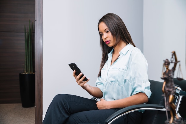 Afroamerikanerfrau mit dem Smartphone, der auf Lehnsessel im Büro sitzt