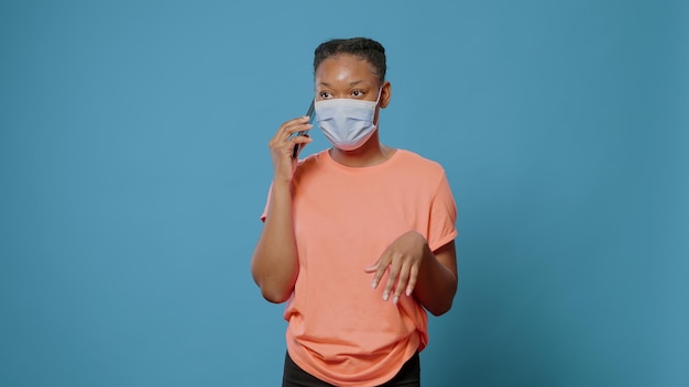 Afroamerikanerin, die sich beim Telefonat unterhält, während sie eine Gesichtsmaske gegen Coronavirus trägt. Junge Person, die während der Covid-19-Pandemie Smartphone verwendet, um mit Freunden im Studio zu sprechen.